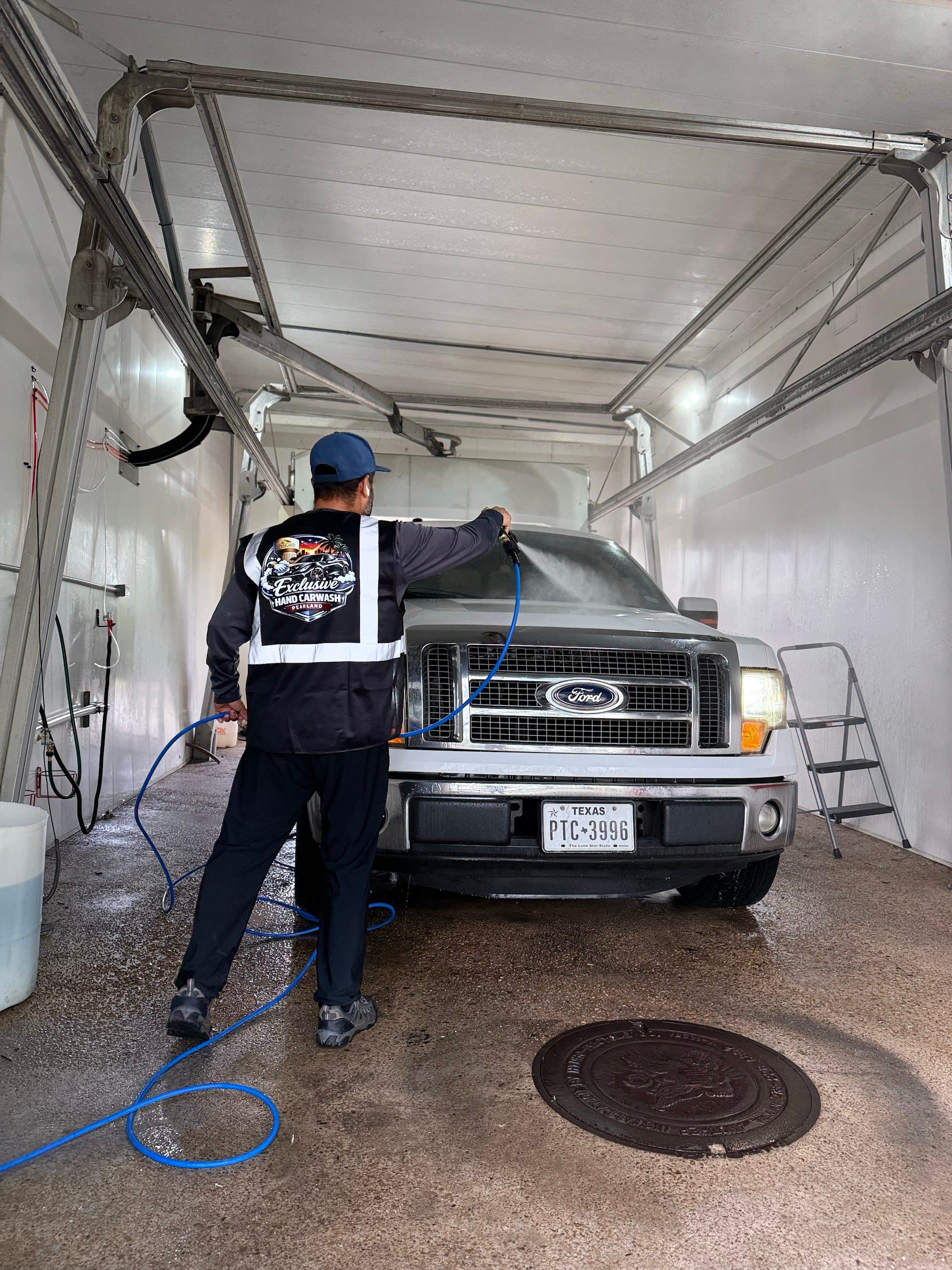 Worker in a branded vest washes a white Ford truck with a high-pressure water hose.