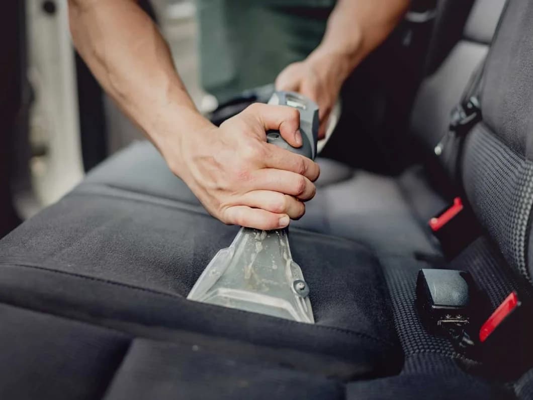 Hands using a professional vacuum extractor to deep clean a black fabric car seat.