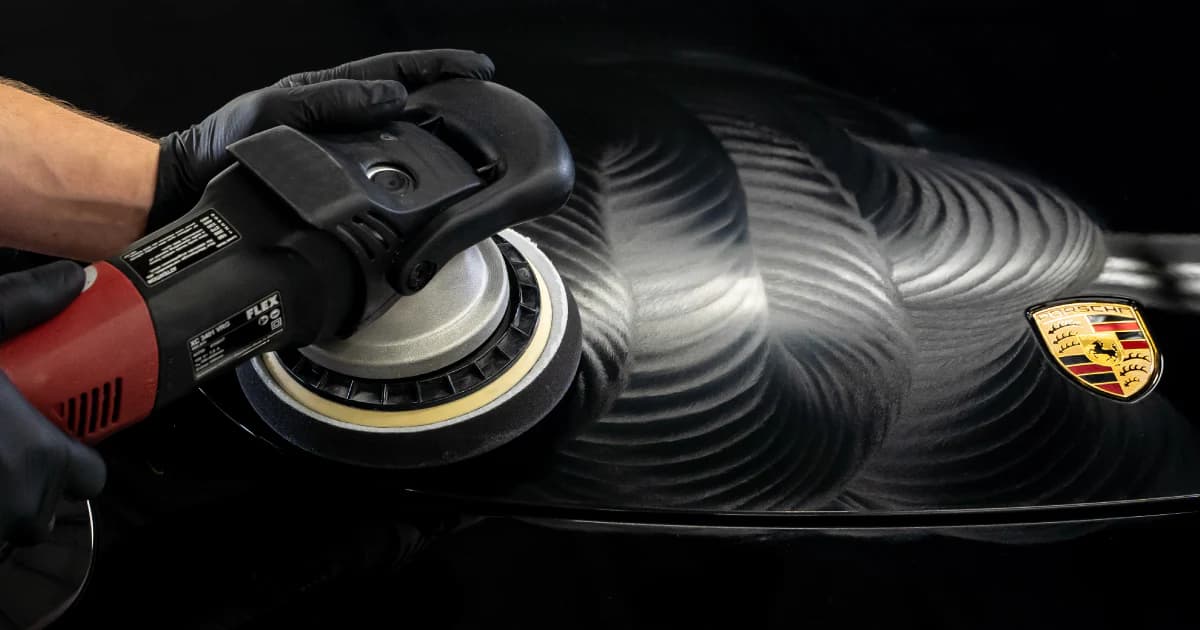 Gloved hands using a power buffer to polish a black Porsche hood with swirl patterns.